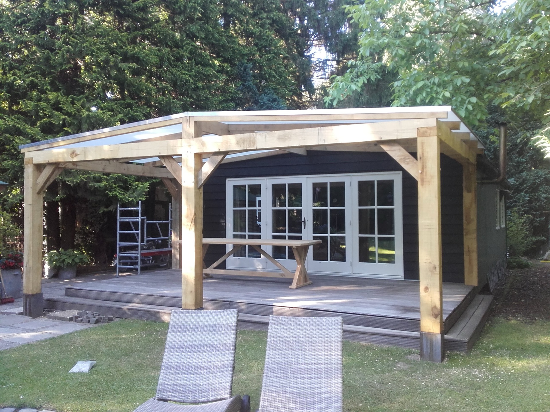 Houten veranda met glazen overkapping tegen een zwart geschilderde woning met witte openslaande deuren