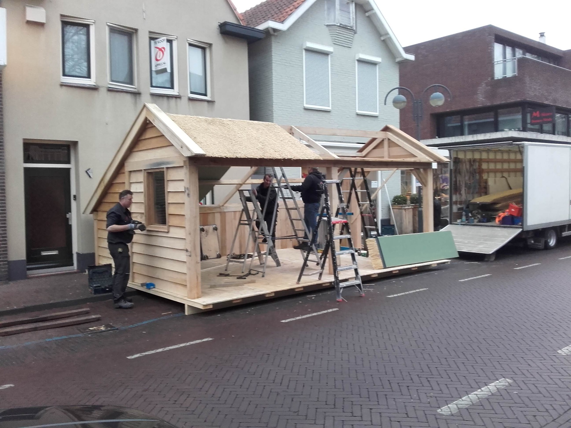 Vakmensen van Trizo monteren op straat een houten tuinhuis met rieten dakbedekking